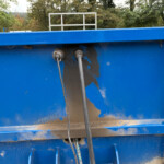 Blue industrial dumpster with two hoses connected to outlets outdoors among trees and grass behind it.