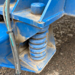 Blue industrial machinery close-up showing stacked spiral springs and a black hose on a dusty gravel surface.