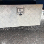 Diamond-plate metal toolbox mounted under a vehicle, with a central latch and small padlock hole visible