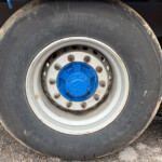 Close-up of a large truck tire with a blue hub and white rim, dirt on the tire surface and ground nearby.