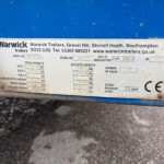 Metal identification plate on a blue trailer showing model, serial number, and manufacture details; dirt around the edge and screws at corners are visible on the plate.