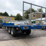 Blue heavy transport trailer in a dealership yard, with a New Holland tractor on it and Russell's/Parts signage in the background.