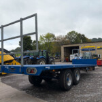 Blue transport trailer and agricultural machinery parked in a yard next to a brick building with New Holland Russell’s signage; several tractors and equipment visible in the background.