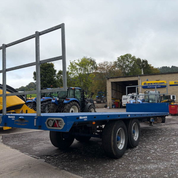 Blue transport trailer and agricultural machinery parked in a yard next to a brick building with New Holland Russell’s signage; several tractors and equipment visible in the background.