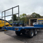 Large outdoor lot with blue farming trailer and yellow tractor on the left; metal hurdle frame in foreground and a workshop with New Holland Agriculture signs in the background.