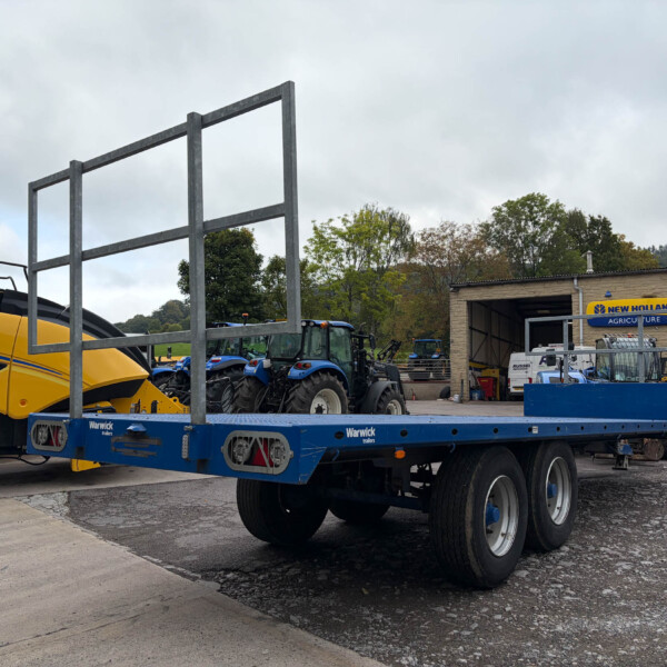 Large outdoor lot with blue farming trailer and yellow tractor on the left; metal hurdle frame in foreground and a workshop with New Holland Agriculture signs in the background.