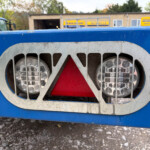 Close-up of a blue vehicle front with round headlights behind a metal grille and a red triangular reflector.
