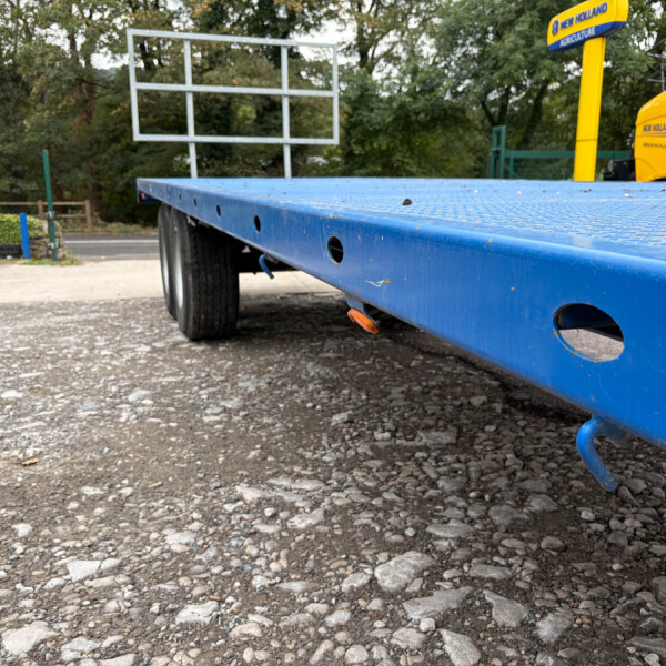 Low-angle view of a blue flatbed trailer on a gravel lot, with a bright yellow truck in the background and trees behind.