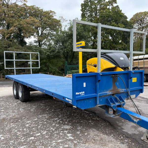 Blue flatbed trailer with metal framing carrying a yellow New Holland agricultural machine, parked in a lot with trees in the background.