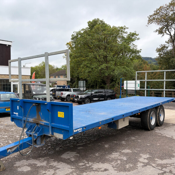 Blue flatbed trailer in a paved car park with a brick building on the left and trees in the background.