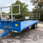 Blue flatbed trailer with a large metal frame structure on a parking lot, with trees and cars in the background