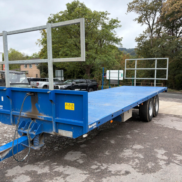 Blue flatbed trailer with a large metal frame structure on a parking lot, with trees and cars in the background