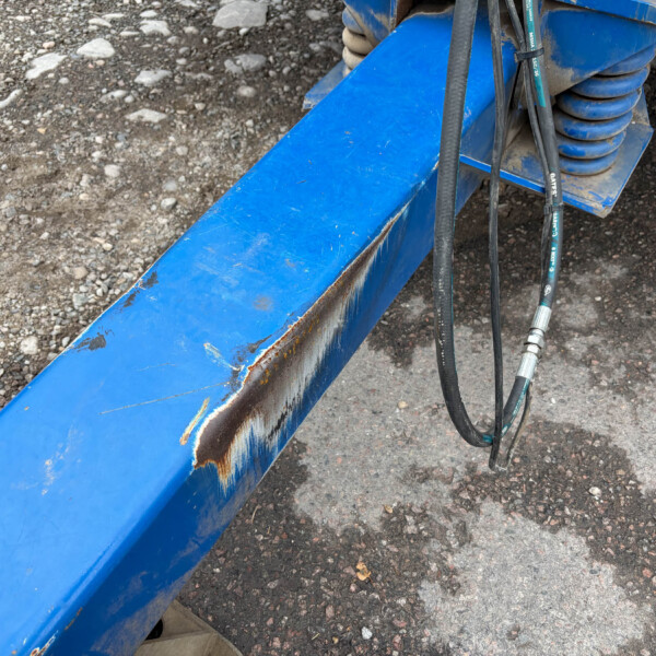 Blue metal beam with rust and scratches at a construction site, with black hydraulic hoses and a coiled spring nearby on gravel ground