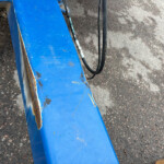 Close-up of a weathered blue metal beam with rust spots, resting on a gravel surface with a black hydraulic hose nearby.